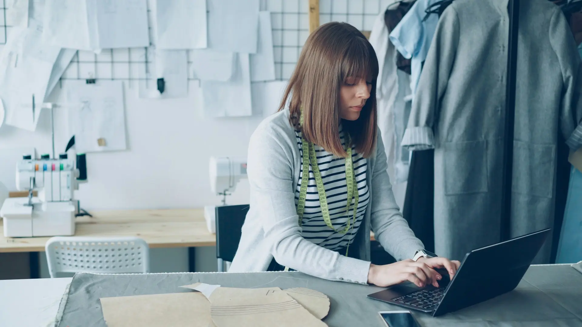 Eine Schneiderin arbeitet in ihrem Atelier am Laptop, im Hintergrund hängen Schnittmuster
