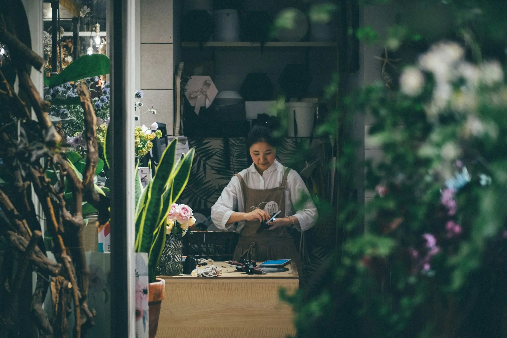 Eine Frau arbeitet an einem Holztresen, umgeben von Pflanzen und Blumen in einem kleinen Laden